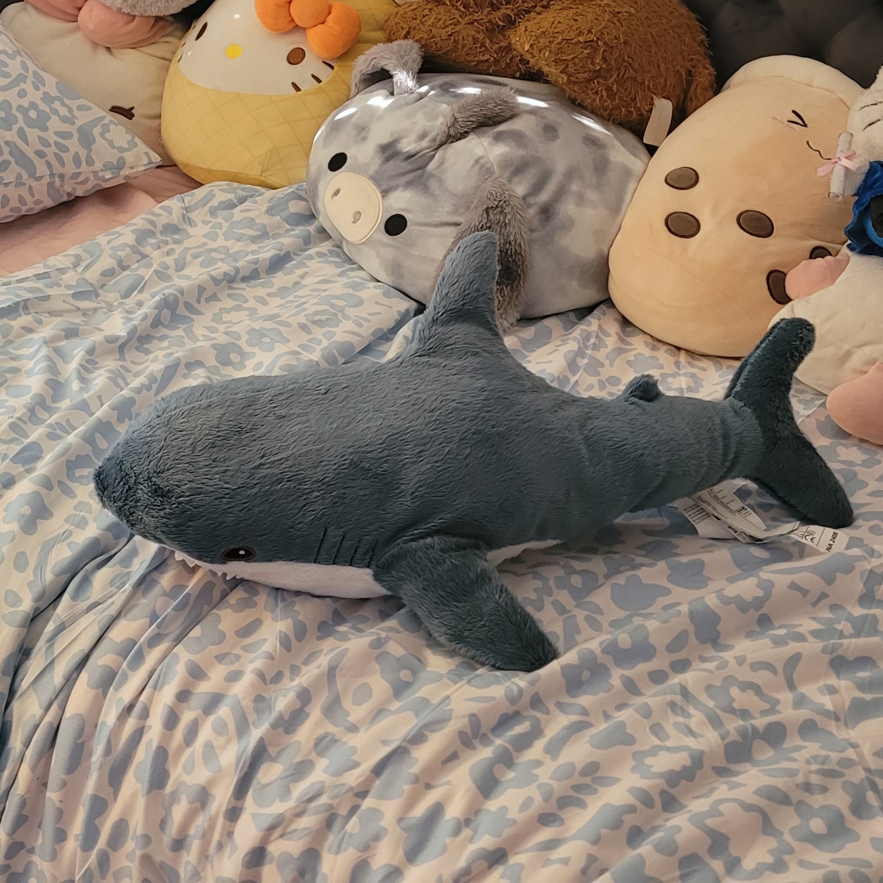 blue shark plushie, called blåhaj, on a bed with a white and blue patterned blanket with a bunch of plushies